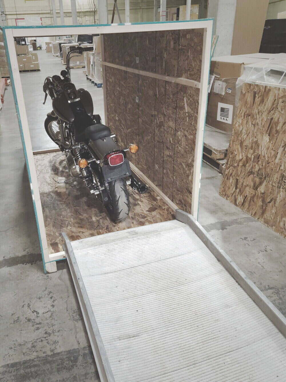A motorcycle strapped upright on the deck of an open custom wood crate, loading ramp down, inside the shop