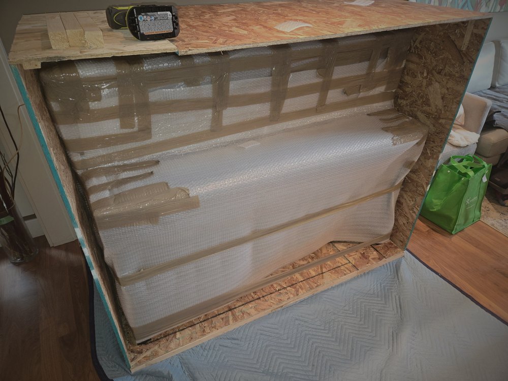 An open wood crate with a large television wrapped in protective film and cross-braced inside